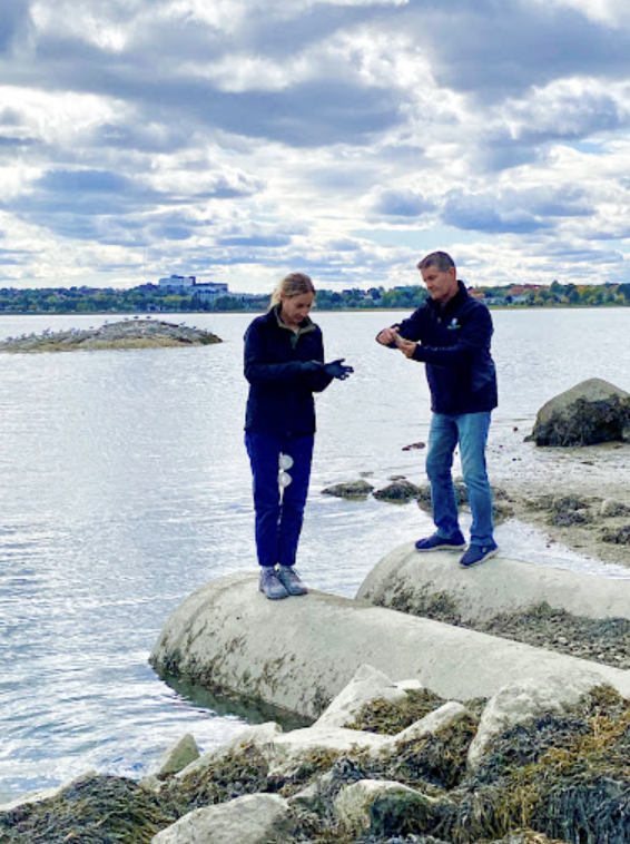 Two people stand on a rocky shoreline collecting data. 